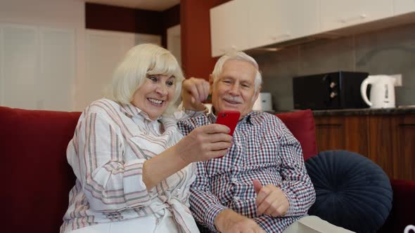 Happy Smiling Senior Couple with Mobile Phone at Home. Resting on Sofa in Cozy Living Room