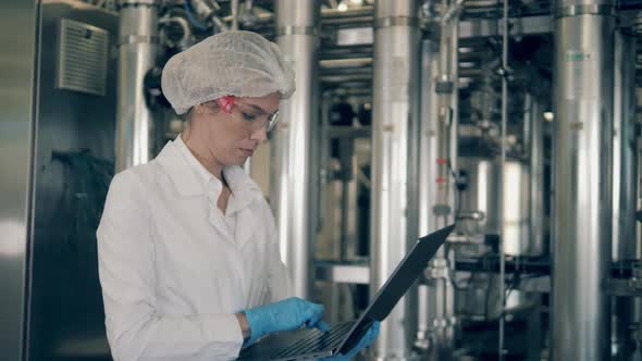 Dairy Plant Worker Types on Laptop. A Woman in a Factory Floor in a White Uniform Controls Quality
