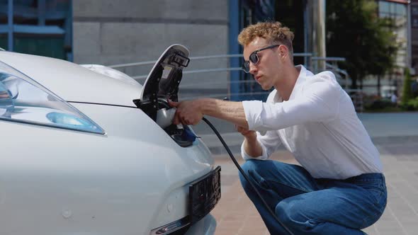 A Young Curly Man Connects an Electric Car to the Charger and Adjusts the Process of Charging the