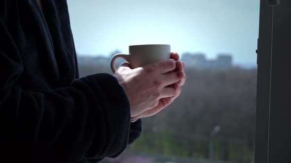 Man Drinks Coffee From Cup in Black Bathrobe at an Open Window in the Morning