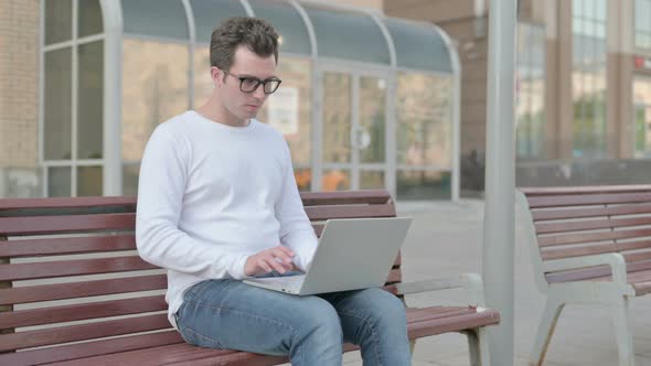 Young Man with Neck Pain Using Laptop While Sitting Outdoor on Bench