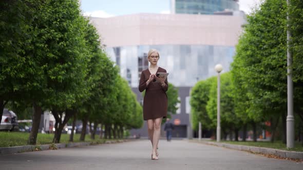 A Business Woman Walks Along an Alley with Trees and Scrolls a Tablet