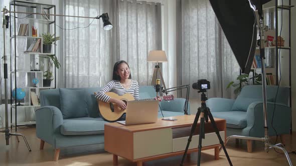 Asian Woman With A Laptop Playing A Guitar And Smiling To Camera While Live Stream At Home