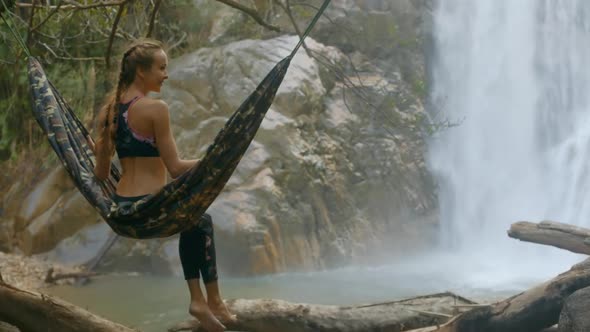 Girl with Plaits Sits on Hammock Against Waterfall