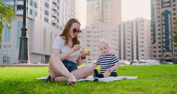 Young Mom with Baby Sitting on the Grass in the Park Eating Lunch