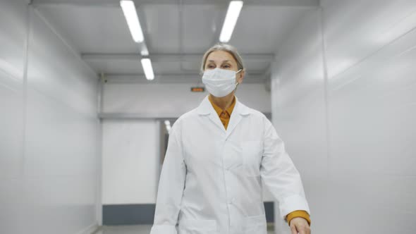 Mature Woman Doctor in White Coat and Safety Mask Walking Along Hospital Corridor