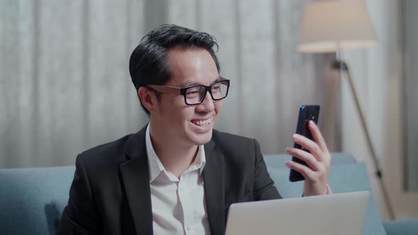 Asian Businessman In Jacket And Shorts Having Video Call On Smartphone While Working With A Laptop