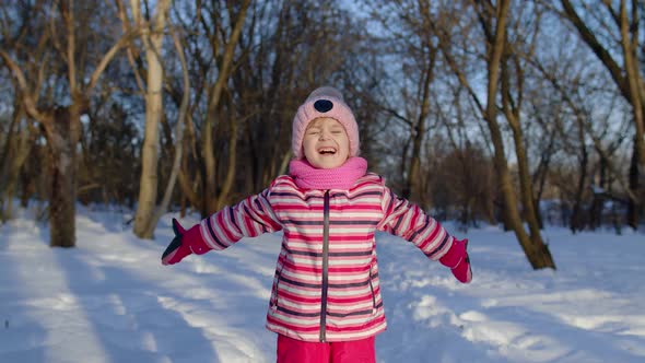 Joyful Little Child Girl Throwing Snow Up Smiling Playing Showing Thumbs Up in Winter Park Forest