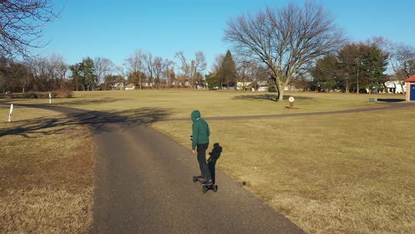 An aerial tracking of a man on an electric skateboard in an empty park on a sunny day. The drone fol