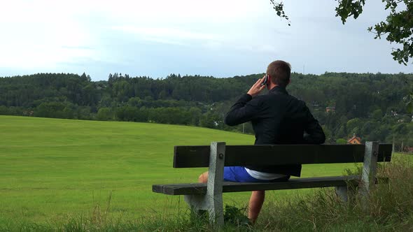 Young Man Sits on the Bench in Nature (Field with Forest) and Calls with Smartphone