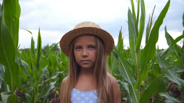 Portrait of Little Serious Girl in Straw Hat Looking Into Camera Against the Background of Corn