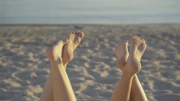 Two Pairs of Slim Young Female Feet at the Background of Sandy Beach and Water. Unrecognizable