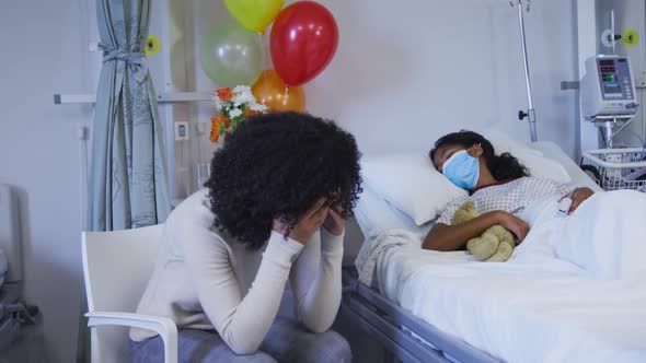 Stressed african american mother sitting besides her her daughter lying on bed at hospital