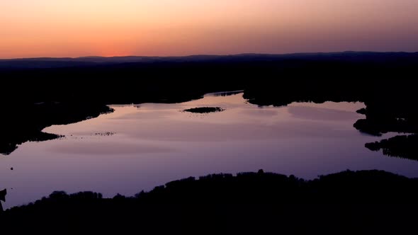 Aerial Over Calm Smooth Lake With Purple Reflected Light Against Orange Sunset Skies Silhouetted By