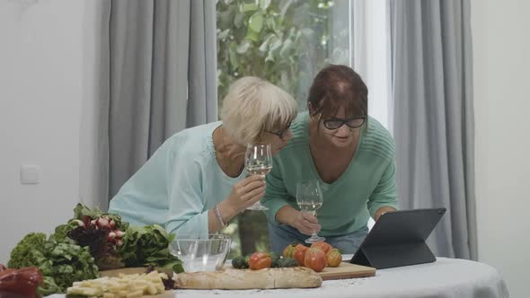 Two Cheerful Smiling Senior Women with Wine Glasses Looking at Tablet Screen and Talking