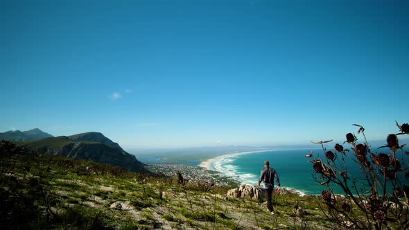 Man walks towards big rock on mountain, and throws hands up into the air triumphantly on it, long st