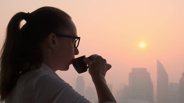 woman drinks tasty coffee while looking out window at evening city at sunset