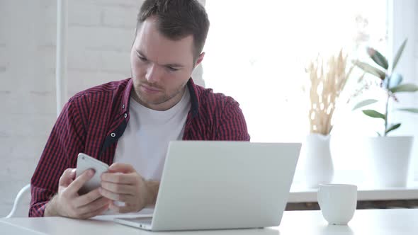 Young Man Using Smartphone While Working on Laptop