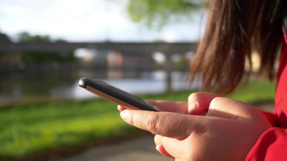 Close up hands of Asian woman wearing coat sitting at the park by the river and typing on her phone