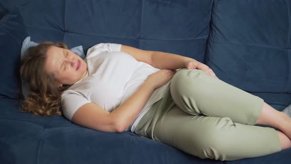 Woman in White Tshirt is Lying on Blue Sofa and Holding Stomach with Hand