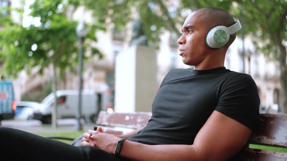 Concentrated bald African man listening music in headphones while sitting on the bench