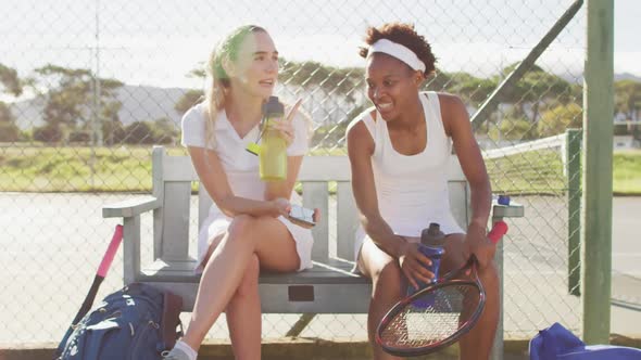 Video of happy diverse female tennis players sitting, talking and resting after match