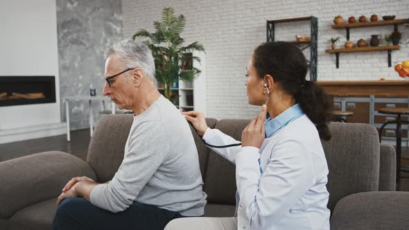 Female Doctor Examining Elderly Male By Phonendoscope Listening to His Lungs Sitting on Couch During