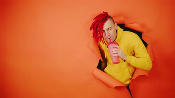 Young Man Drinking Through Soft Drink Straw in Hole of Orange Background