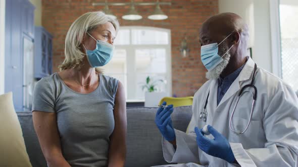 Happy senior diverse woman and doctor wearing face masks in living room sitting on sofa, vaccinating