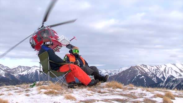 Two men sit on the top of a mountain as a helicopter drops off supplies.