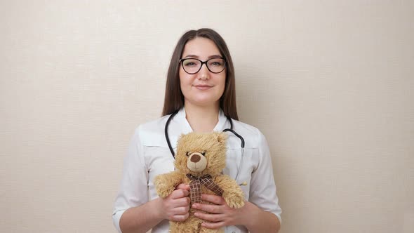 Woman Pediatrician with Glasses Holds Teddy Bear on Beige