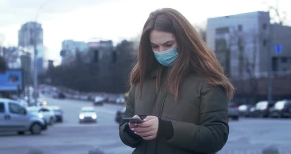 Portrait of Worried Woman Standing on City Street in Protective Mask and Using Smartphone. Busy