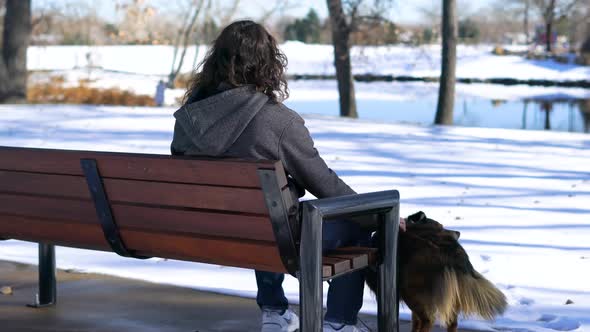 Man with dark long hair sitting at a bench petting his dog in Winter at a park