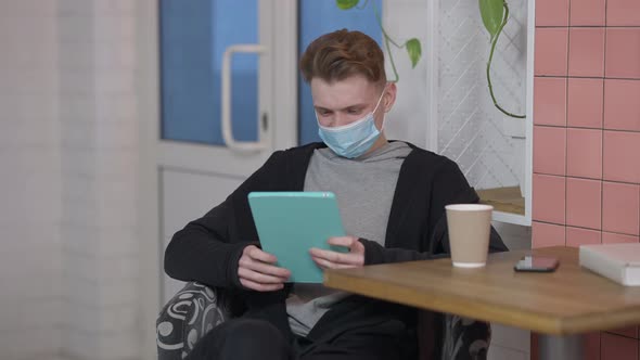 Portrait of Young Positive Man in Covid Face Mask Surfing Social Media on Tablet Smiling
