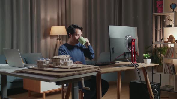 Asian Male Engineer With The House Model Drinking Coffee While Working On A Desktop At Home