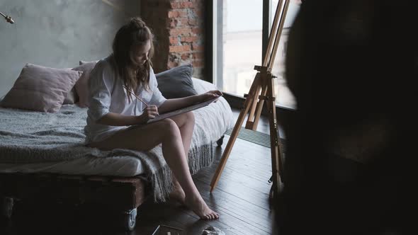 Girl Artist Paints a Picture of Sitting on the Bed in the Shirt. Painter Works in a Home Studio in