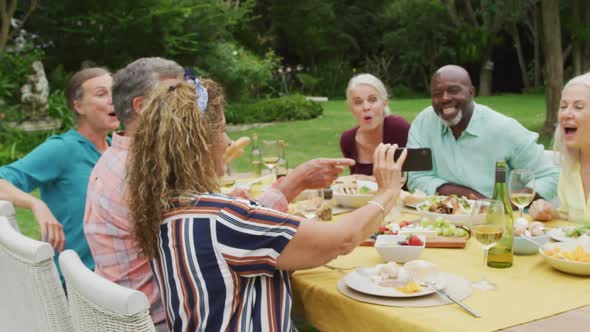 Animation of diverse happy senior female and male friends eating lunch in garden, using smartphone