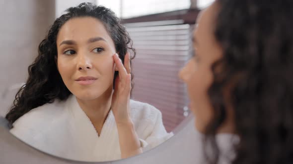 Young Attractive Woman in Bathrobe Looks in Mirror in Bathroom Touches Moisturized Face After