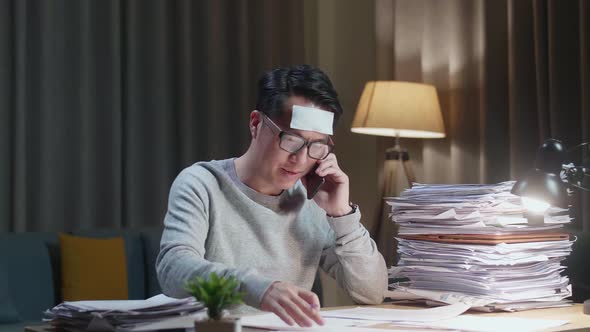 Sick Asian Man Talking On Phone And Taking Note While Working Hard With Documents At Home