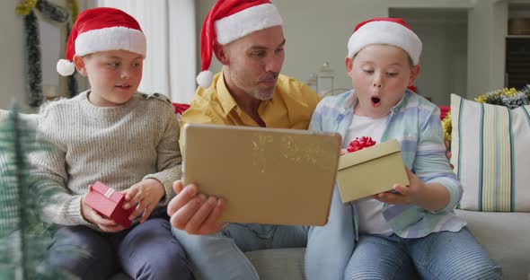 Caucasian father and two sons opening gifts having a videocall on digital tablet during christmas