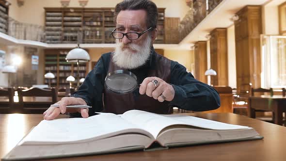 Wise Male Pensioner with Beard in Glasses Sitting in the Library and Flipping Through Book Pages