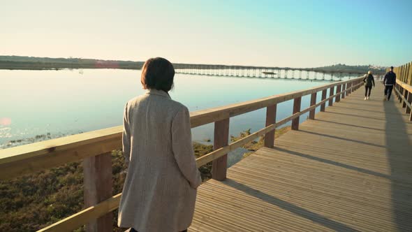 Two Females Walking on a Boardwalk Near a River Shore with People Ahead