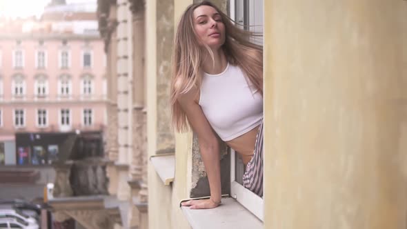 Girl in Pajamas on the Windowsill of a Large Old Building Window Looks Out the Window in the Morning