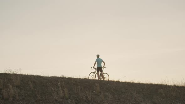 A Man with a Bicycle Stands on the Mountain Raises His Hands To the Top Looking at the Sunset, a