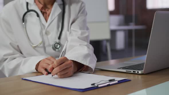 Caucasian female doctor wearing mask sitting at desk in meeting room making notes