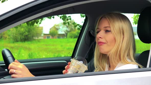 Young Attractive Blond Woman Sits in the Car and Drinks Ice Coffee in Cup