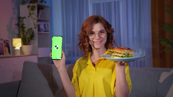 Woman in Glasses with Burger and Mobile Phone with Green Chroma Key in Hands Smiles and Watches at