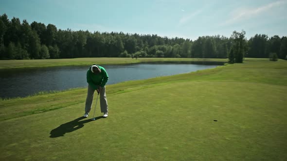 A Golfer in a White Cap and a Green Hoodie Hits a Golf Ball with a Stick and Rolls It Into the Hole