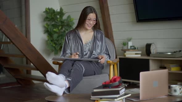 Motivated Young Asian Woman in Pajamas and Eyeglasses Discussing Business Idea Online in Video Chat