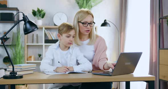 Mother in Glasses which Sitting at Home Desk with Her Handsome Son and Helping Him